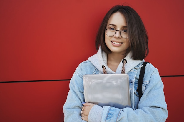 Close-up Outdoor Portret Europejskiej Studentki W Okularach, Atrakcyjna Dziewczyna Stojąca W Pobliżu Czerwonej ściany, Trzymając Laptopa W Rękach, Uśmiechnięta Kamera Z Wesołym Zrelaksowanym Wyrazem.