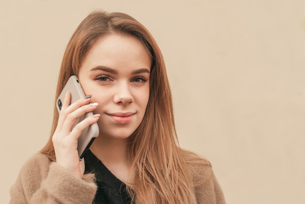 Close-up portret blondynki dzwoni na telefon i patrzy w kamerę na tle beżowej ściany, patrząc w kamerę