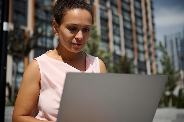 Close-up portret kobiety w średnim wieku Latynoska lub Afroamerykanka, biznes dama, kierownik biura, pracownik, pracownik w swobodnym stroju pracującym na laptopie na tle wysokich budynków