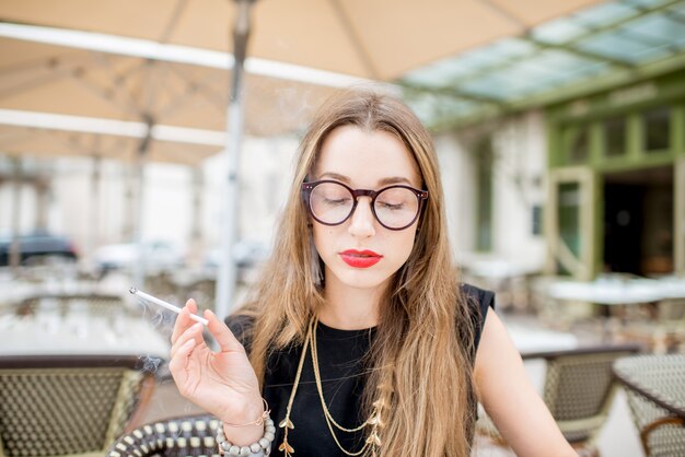 Close-up portret młodej i eleganckiej kobiety palącej papierosa siedzącej na świeżym powietrzu na tarasie typowej francuskiej kawiarni we Francji