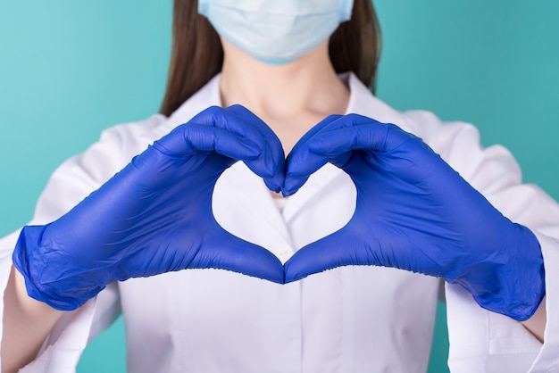 Close-up przycięte zdjęcie kobiety lekarz w białym płaszczu jednolite gumowe lateksowe niebieskie rękawiczki, co pokazuje serce z rękami na białym tle turkusowy niebieski turkusowy