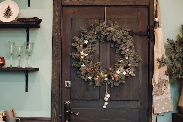 Close-up z jodły Boże Narodzenie wieniec z dekoracjami wiszącymi na ciemnobrązowe drewniane drzwi wejściowe domu. Ozdobiony wieniec bożonarodzeniowy na drzwiach wejściowych.