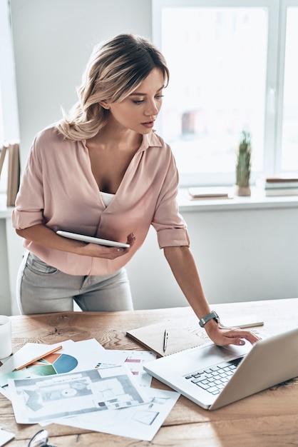 Co jeśli? Piękna młoda kobieta w eleganckim stroju casual pracuje przy laptopie stojąc w nowoczesnym biurze