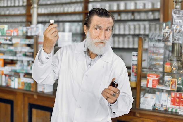 Zdjęcie co jest lepsze portret doświadczonego mężczyzny starszego farmaceuty pracującego w starej aptece trzymającego w rękach dwie butelki z lekami