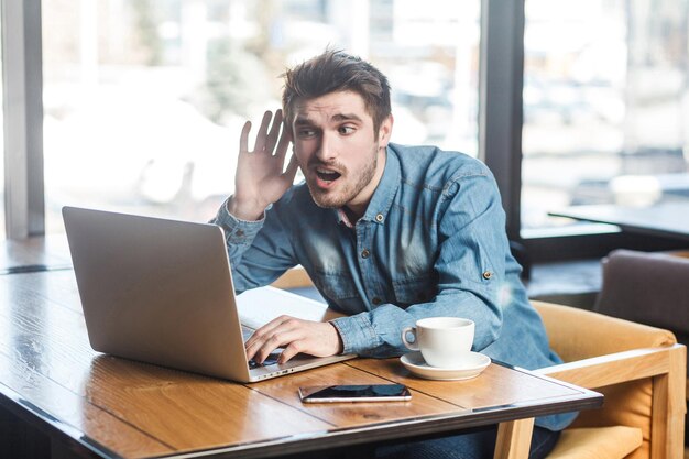 Co mówisz? Widok z boku portret interesujący brodatego młodego freelancera w niebieskiej koszuli dżinsowej siedzi w kawiarni i nawiązuje wideorozmowę na laptopie, chude rękę na uchu, która nie słyszy towarzysza.