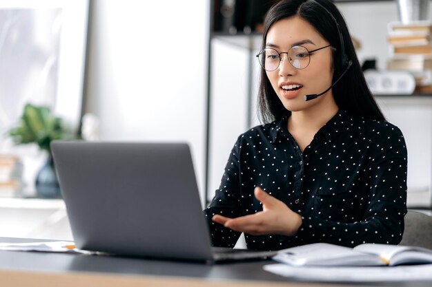 Confident pretty smart asian girl with glasses and headphones freelancer call center operator sitting at desk in office talks to client or employee via video communication gesturing with her hand
