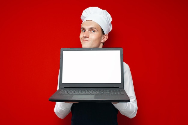 Cook pokazuje pusty ekran laptopa na czerwonym, odizolowanym tle Facet w mundurze szefa kuchni