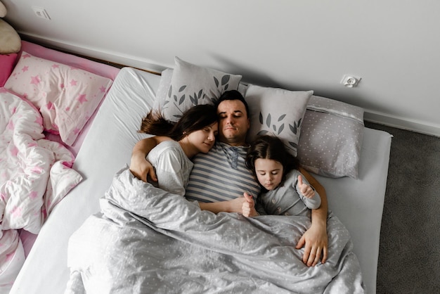 Córka z rodzicami w łóżku, tata delikatnie całuje żonę i córkę, mocne rodzinne uściski, rodzinny weekend w łóżku, śpiąca rodzina, córka śpi z młodymi rodzicami w łóżku