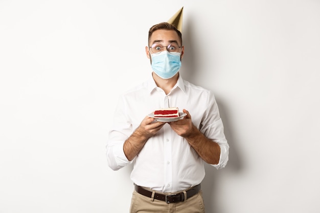 Covid-19, dystans społeczny i świętowanie. Zaskoczony urodzinowy facet trzyma tort urodzinowy, ma na sobie maskę na twarz z koronawirusa, białe tło.