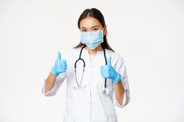 Covid-19 i koncepcja pracowników medycznych. Uśmiechnięta azjatycka kobieta lekarz w masce na twarz i gumowych rękawiczkach pokazuje kciuk w górę, białe tło.
