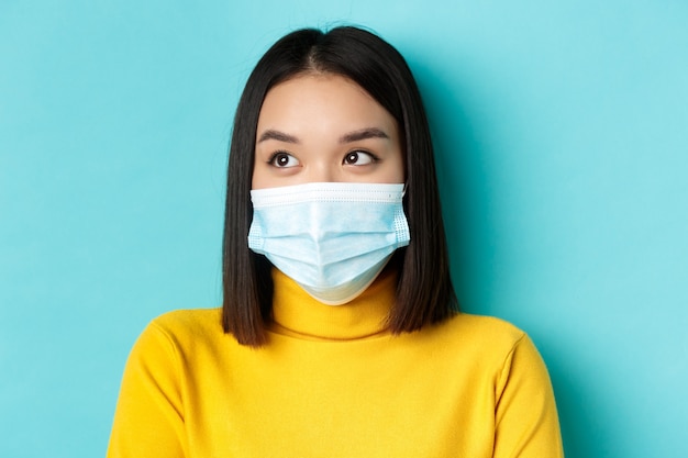 Covid-19, pojęcie dystansu społecznego i pandemii. Headshot ślicznej azjatyckiej kobiety z krótkimi ciemnymi włosami i maską medyczną, patrzącą w lewo, stojącą na niebieskim tle