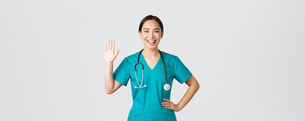 Covid-19, pracownicy służby zdrowia i koncepcja zapobiegania wirusom. Przyjazna uśmiechnięta azjatycka lekarka, lekarz w fartuchu machający ręką, aby powiedzieć cześć, cześć, pozdrawiam pacjenta, miło cię widzieć.