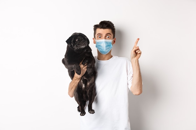 Covid-19, zwierzęta i koncepcja kwarantanny. Młody człowiek w masce medycznej, trzymając ładny czarny mops, pies patrząc w prawo i właściciel wskazujący na logo, biały