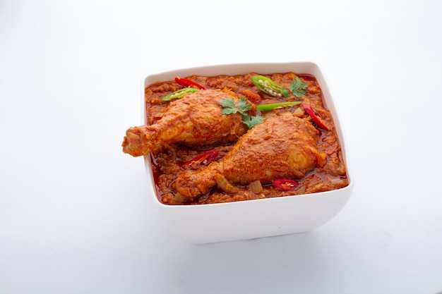 Curry z kurczaka lub masala, pikantne, czerwonawe danie z kawałka nogi kurczaka przyozdobione liściem kolendry i świeżym zielonym chili, które jest ułożone w białej ceramicznej misce z białym tłem, na białym tle.