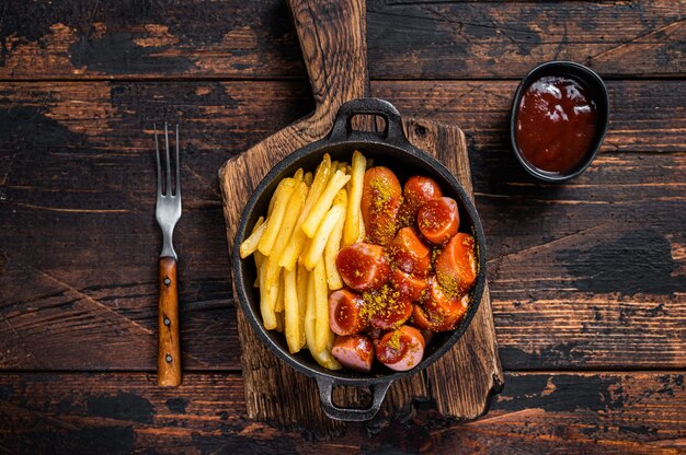 Zdjęcie currywurst street food, przyprawa curry na wursts podawana na patelni z frytkami. ciemny drewniany stół. widok z góry.