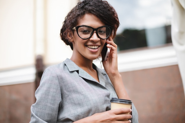 Cute African American girl w okularach stojących na ulicy i rozmawia przez telefon z kawą w ręce