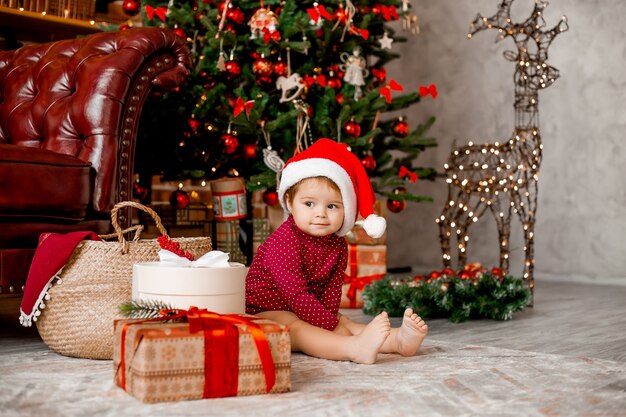 Cute baby Santa siedzi w domu w pobliżu choinki z prezentami