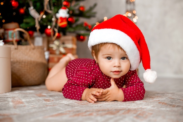 Cute baby Santa siedzi w domu w pobliżu choinki z prezentami