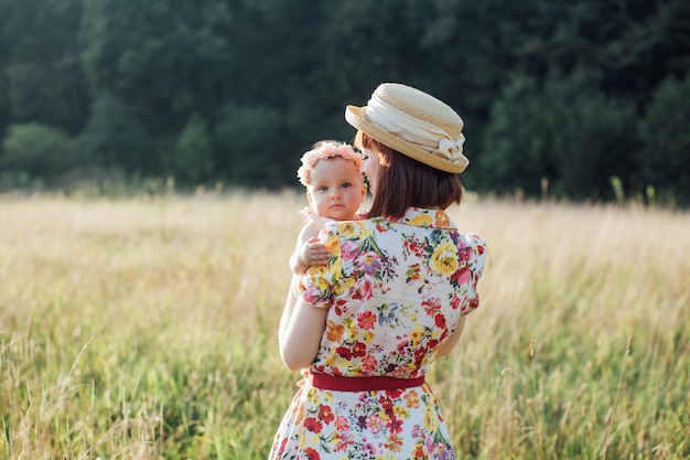 Cute dziewczynka i mama przytulają się w polu lato. Widok z tyłu ładna kobieta matka trzyma swoje dziecko na rękach. Mała córka patrzy na aparat. Szczęśliwa rodzina, mama i córka na polu