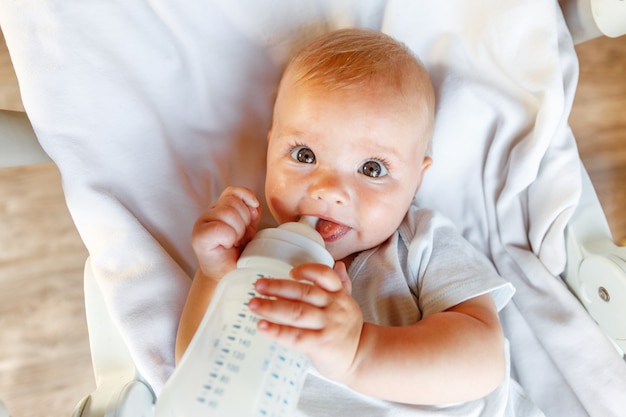 Cute dziewczynka noworodka pije mleko z butelki i patrząc na kamery na białym tle. Niemowlę niemowlę ssanie odżywianie mleka w pozycji leżącej na łóżeczku w domu. Koncepcja szczęśliwego dziecka macierzyństwa.