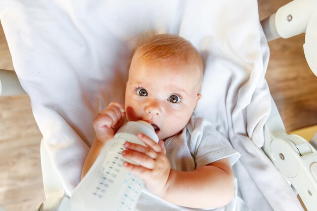 Cute dziewczynka noworodka pije mleko z butelki i patrząc na kamery na białym tle. Niemowlę niemowlę ssanie odżywianie mleka w pozycji leżącej na łóżeczku w domu. Koncepcja szczęśliwego dziecka macierzyństwa.