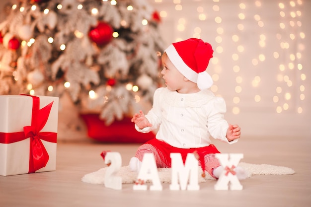 Cute dziewczynka sobie santa claus siedzi na podłodze z białymi drewnianymi literami Boże Narodzenie w pokoju. Odwracając wzrok. Sezon wakacyjny.
