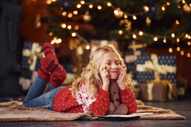 Cute dziewczynka w czerwony świąteczny sweter w pozycji leżącej z misiem pomieszczeniu z okazji nowego roku.
