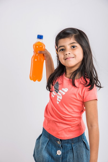 Cute indyjskich lub azjatyckich dziewczynka z butelką PET zimnego napoju lub soku owocowego pomarańczowy lub mango, picie lub trzymając stojąc na białym tle na niebieskim lub białym tle.