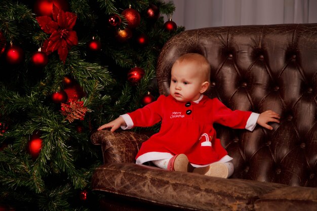 Cute jednoroczna dziewczyna w stroju Świętego Mikołaja na kanapie na tle choinki zdobione zabawki. Dziecko w dekoracjach z pudełkami na prezenty. Koncepcja przytulnego domu obchody szczęśliwego nowego roku. Skopiuj miejsce