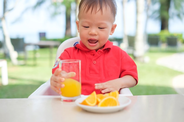 Cute little azjatyckich toddler chłopiec dziecko siedzi w wysokim krześle trzymając i pije smaczny sok pomarańczowy zimny napój w szklance podczas śniadania