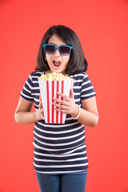 Cute little Indian girl jedzenie popcornu podczas noszenia okularów przeciwsłonecznych lub okularów 3D w teatrze. Stojąc na białym tle nad kolorowym tłem