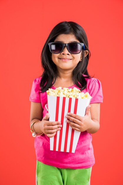 Cute little Indian girl jedzenie popcornu podczas noszenia okularów przeciwsłonecznych lub okularów 3D w teatrze. Stojąc na białym tle nad kolorowym tłem