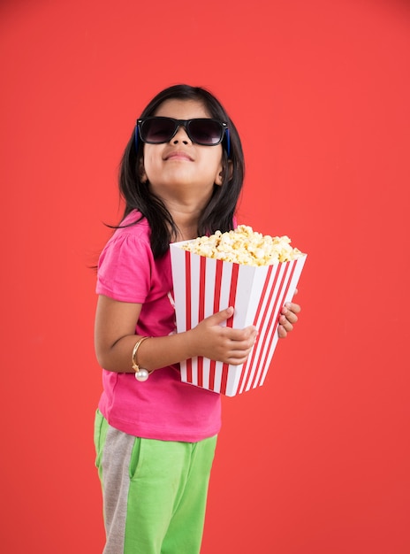 Cute little Indian girl jedzenie popcornu podczas noszenia okularów przeciwsłonecznych lub okularów 3D w teatrze. Stojąc na białym tle nad kolorowym tłem