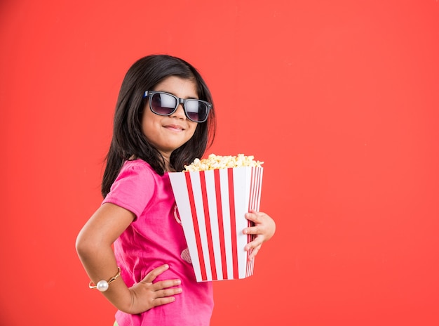 Cute little Indian girl jedzenie popcornu podczas noszenia okularów przeciwsłonecznych lub okularów 3D w teatrze. Stojąc na białym tle nad kolorowym tłem