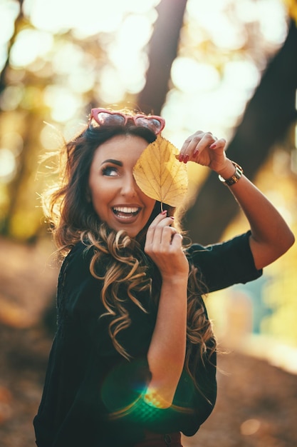 Cute młoda kobieta cieszy się w słonecznym lesie w jesiennych kolorach. Trzyma złoty liść i odwraca wzrok.