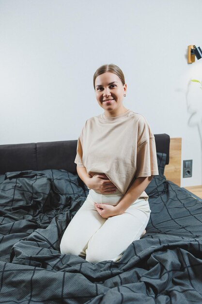 Cute młoda kobieta w ciąży siedzi na łóżku z szarymi prześcieradłami w ubraniach dorywczo Cute kobieta w ciąży w domu