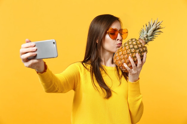 Cute młoda kobieta w okulary trzymać, całując owoce ananasa robi selfie strzał na telefon komórkowy na białym tle na żółtym pomarańczowym tle. Ludzie żywy styl życia zrelaksować koncepcję wakacji. Makieta miejsca na kopię