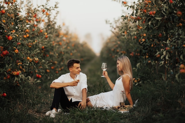 Cute para na pikniku siedząc na zielonej trawie w sadzie jabłkowym. Romantyczna para opiekania winem, siedząc razem na pikniku.