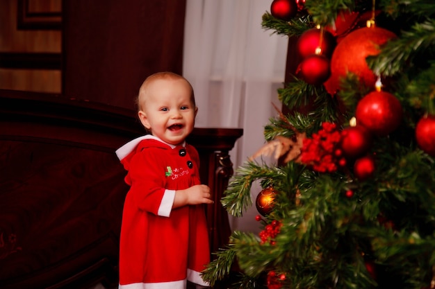 Cute roczna dziewczynka w stroju Świętego Mikołaja na tle choinki ozdobiony zabawkami. Dziecko w dekoracjach z pudełkami na prezenty. Koncepcja przytulnego domu obchody szczęśliwego nowego roku. Skopiuj miejsce