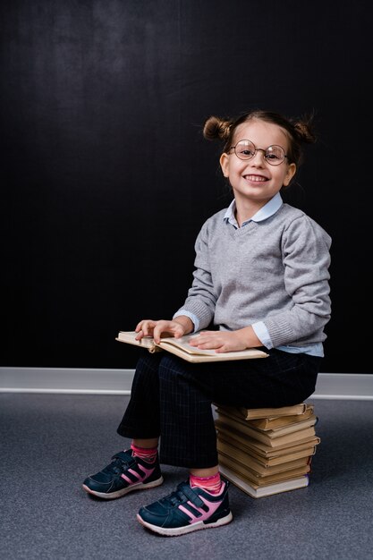 Cute Uśmiechnięta Uczennica Siedzi Na Stosie Książek Podczas Czytania Jednego Z Nich Na Tablicy W Izolacji