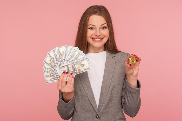 Cyfrowe pieniądze bitcoin Portret szczęśliwa optymistyczna kobieta w eleganckiej marynarce trzymającej banknoty dolarowe i złotą monetę btc uśmiecha się do aparatu studio strzał na białym tle na różowym tle