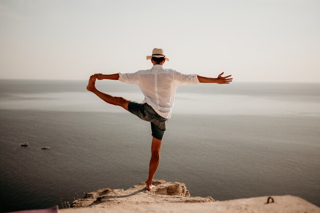 Cyfrowy nomada mężczyzna w kapeluszu biznesmen ćwiczy jogę na skałach nad morzem o zachodzie słońca robi