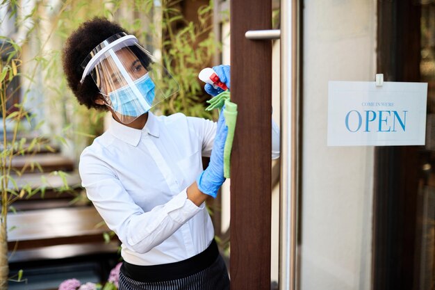 Czarna kelnerka czyści drzwi wejściowe podczas ponownego otwierania kawiarni podczas epidemii COVID19