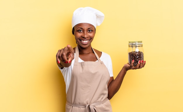 Czarna kobieta kucharz afro, wskazując na aparat z zadowolonym, pewnym siebie, przyjaznym uśmiechem, wybierając Ciebie. koncepcja ziaren kawy
