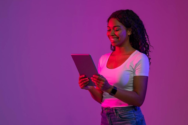 Czarna kobieta stojąca w neonowym świetle i przeglądająca internet na cyfrowym tablecie