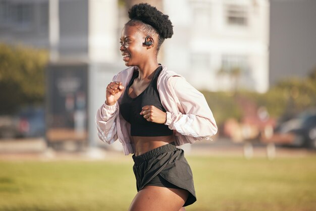 Czarna kobieta uśmiecha się i biega na świeżym powietrzu ze słuchawkami muzycznymi do słuchania radia lub podcastu Sporty fitness i szczęśliwy afrykański sportowiec trenują i ćwiczą dla zdrowia, odnowy biologicznej lub cardio