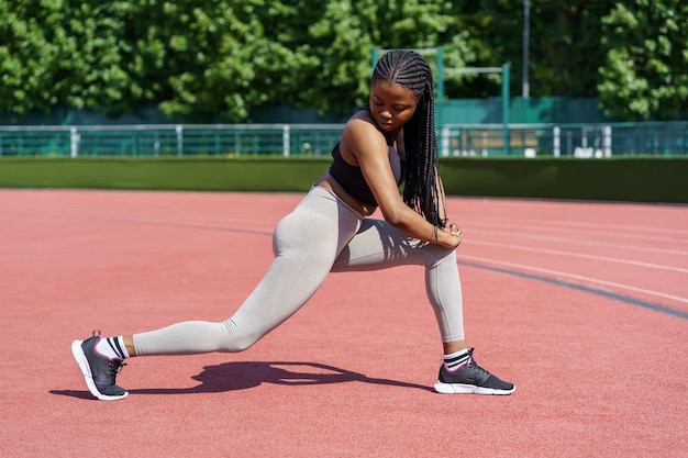 Czarna kobieta w stroju sportowym pompuje mięśnie pośladkowe na boisku sportowym w pobliżu parku miejskiego z bujnymi drzewami