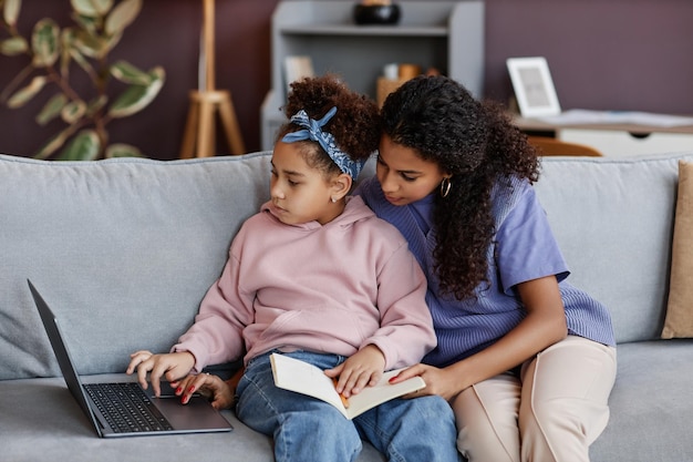 Czarna młoda kobieta uczy córkę za pomocą laptopa siedząc razem na kanapie