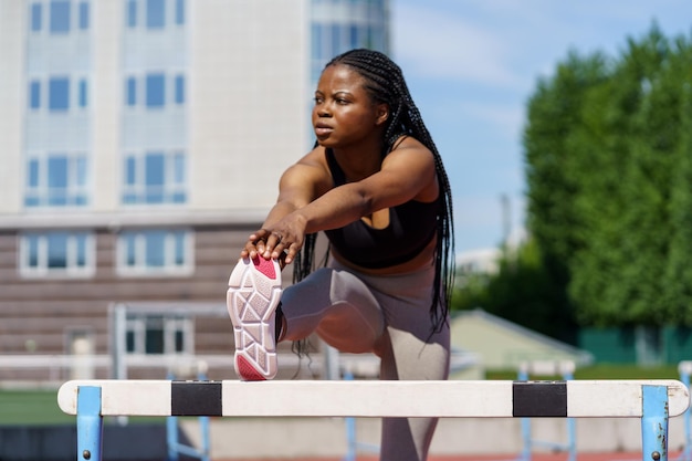 Czarna młoda kobieta z długimi warkoczami rozciąga nogi i ciało za pomocą bariery przez płotki na boisku sportowym