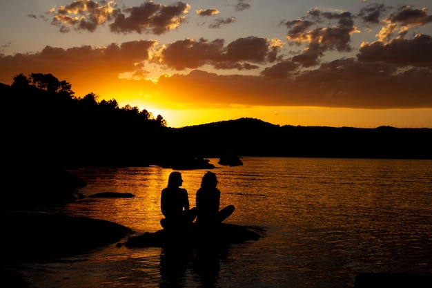 Czarna sylwetka dwóch przyjaciół siedzących nad jeziorem w złotym zachodzie słońca. Skopiuj miejsce.